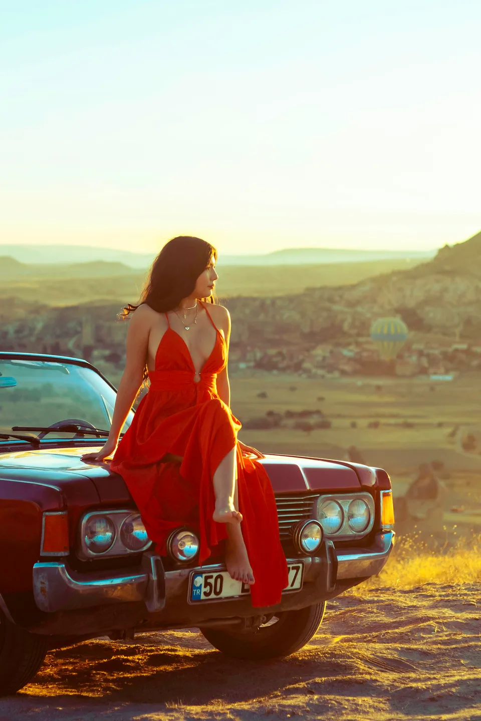 A woman in a red dress on a vintage car above the Cappadocia valleys